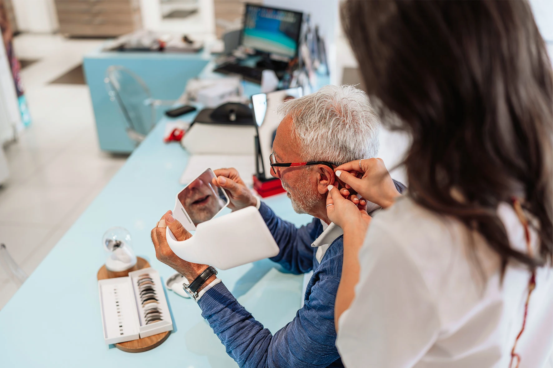 Optikerin passt einem Senior eine Brille an, Spiegel und Brillengestell am Tisch
